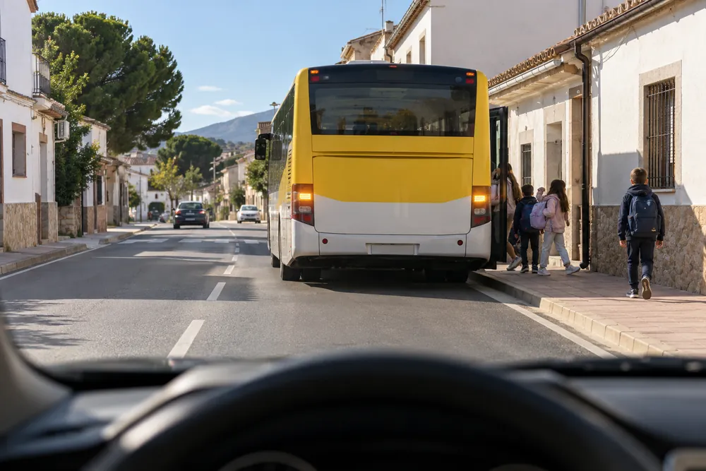Autobús escolar detenido junto a la acera mientras bajan niños, visto desde el coche que se acerca.