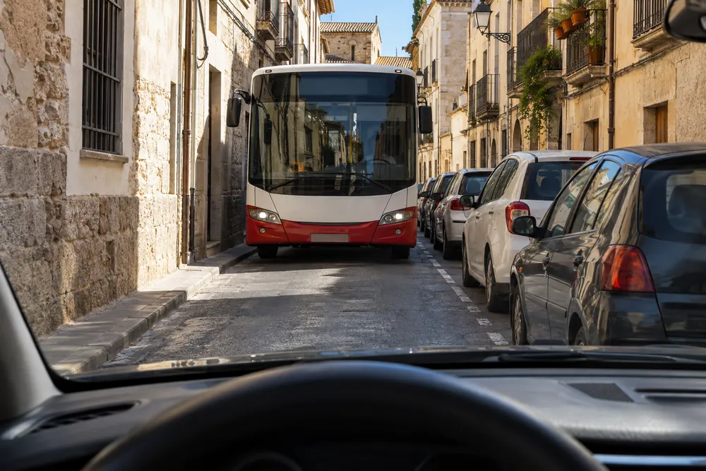 Autobús urbano que se acerca de frente por una calle estrecha donde no cabe junto al turismo.