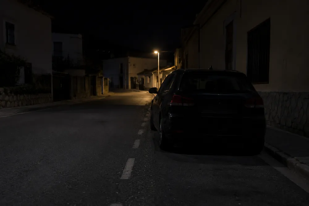 Calle mal iluminada de noche con un turismo estacionado junto al bordillo.