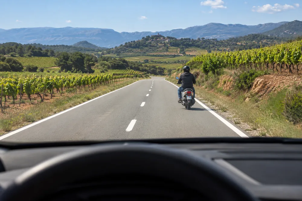 Ciclomotor circulando por el borde derecho de una carretera convencional, visto desde el turismo que se acerca por detrás.