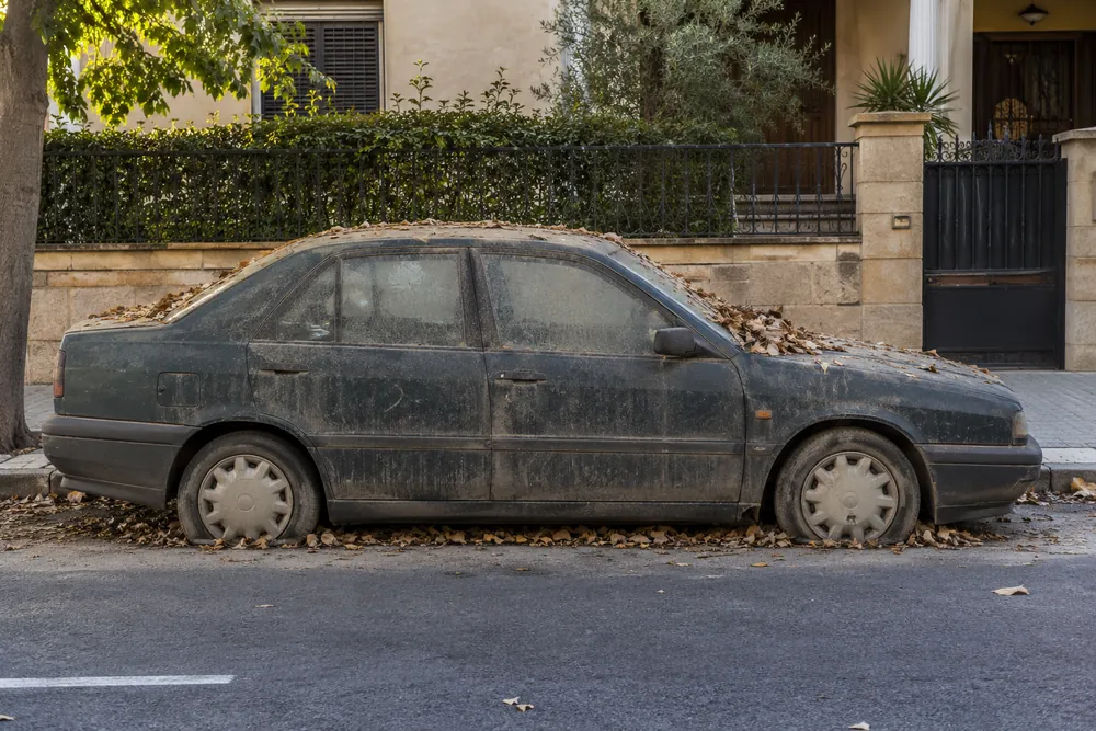 Turismo cubierto de polvo y hojas, con las ruedas desinfladas, estacionado en una calle.