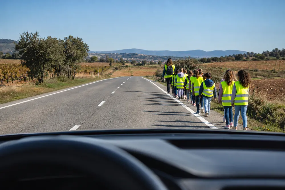 Grupo de escolares caminando en fila por el arcén de una carretera con un monitor.