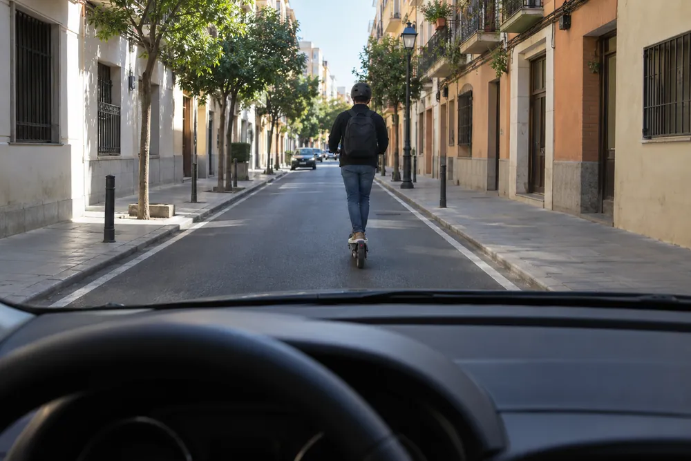 Patinete eléctrico circulando por el centro del carril delante de un turismo en una calle.
