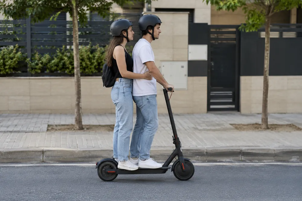 Dos jóvenes adultos sobre un único patinete eléctrico de una plaza.