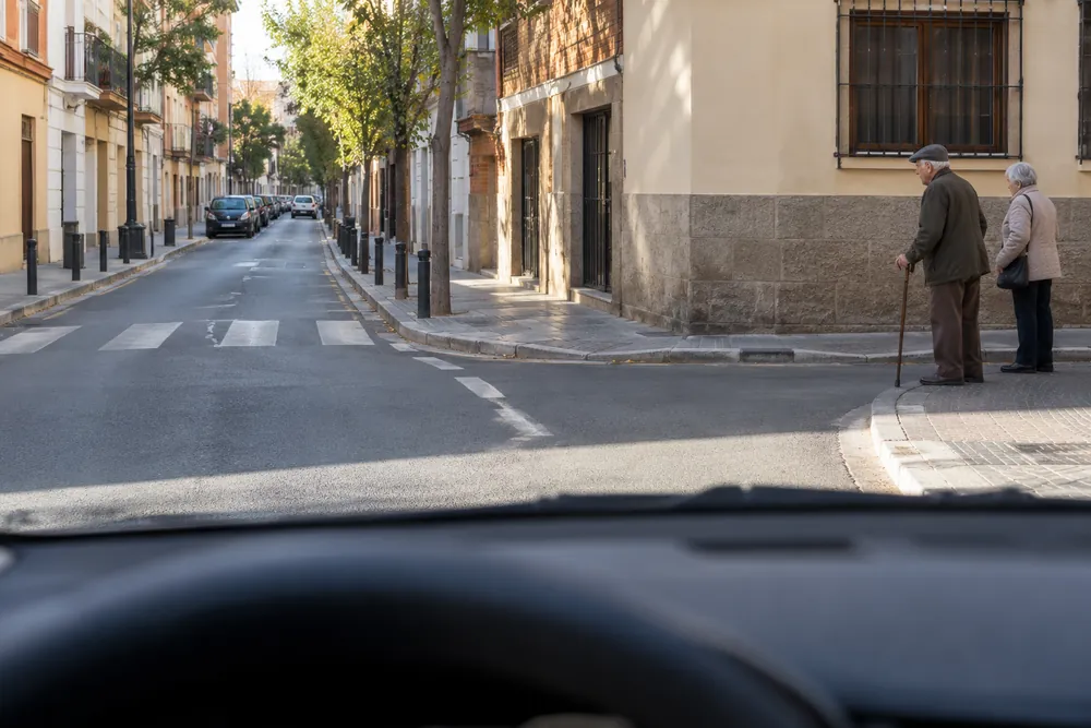 Dos personas mayores junto al bordillo de un cruce urbano.