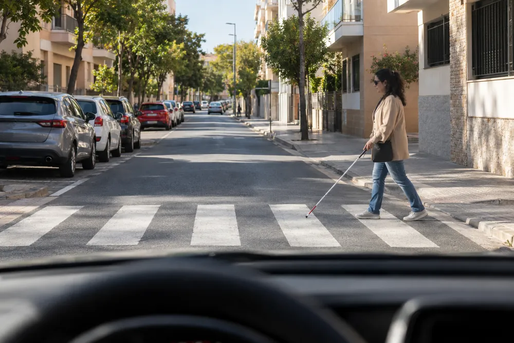 Persona con bastón blanco cruzando por un paso de cebra, vista desde el coche que se aproxima.