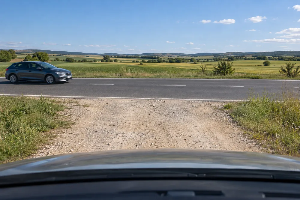 Turismo detenido al final de un camino de tierra, a punto de incorporarse a una carretera asfaltada por la que circula otro coche.