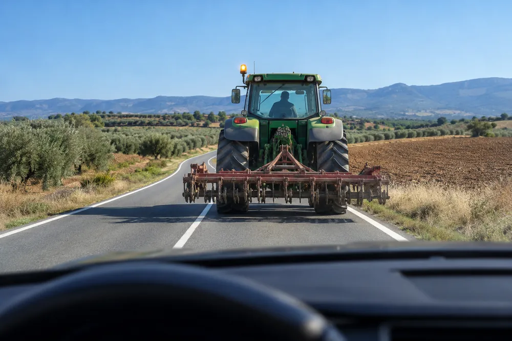 Tractor con apero circulando despacio por una carretera convencional, con la luz amarilla giratoria encendida.